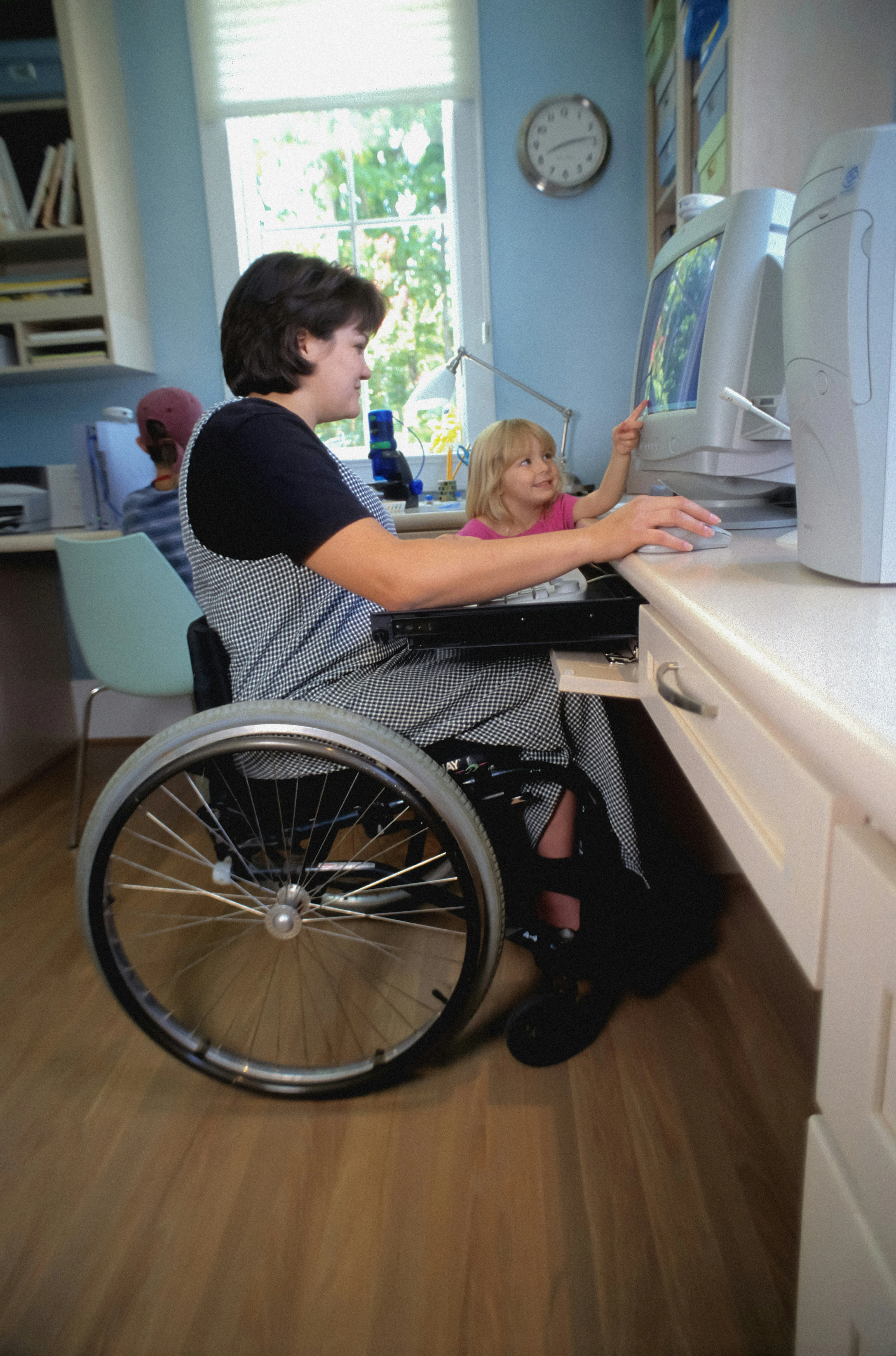 Frau im Rollstuhl bedient einen Computer, ein Kind steht daneben